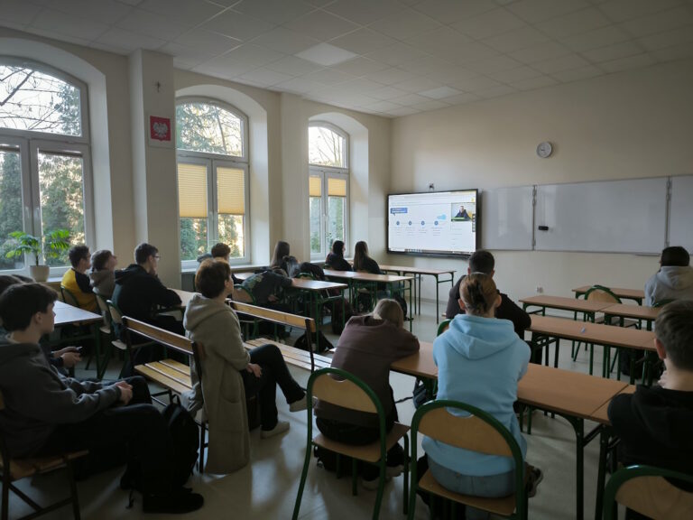 Uczniowie siedzący w szkolnych ławkach i oglądający prezentację na interaktywnym monitorze