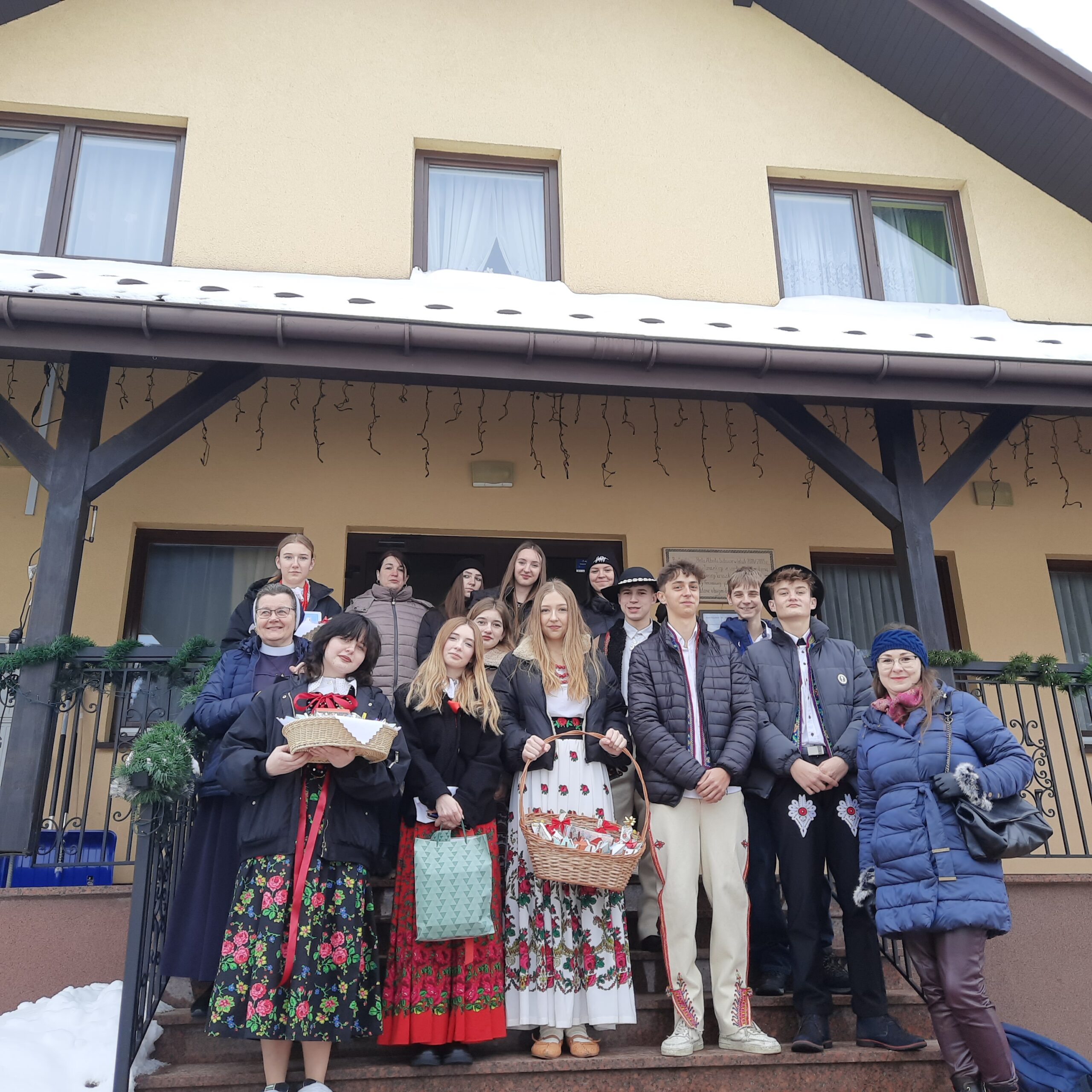 Młodzież w strojach regionalnych wraz z opiekunami stojąca na schodach przed Przytuliskiem świętego Brata Alberta w Grywałdzie.